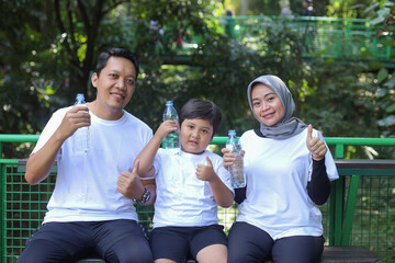 Happy Muslim family holding bottle with water while resting after sport workout outdoors in the public park. Parents and son smiling and giving thumbs up gesture.