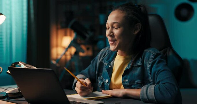 Portrait of a smiling student studying diligently in front of a laptop, taking notes with pencil in notebook, remote learning, preparation for exams, tests, distance learning tutoring.