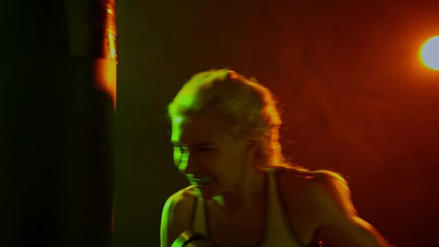 Sportive woman, female professional boxer practices punch on punching bag in dark smoky gym in neon light. Sport, action, power, energy concept.
