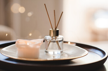 Home liquid scented fragrance in glass bottle with bamboo sticks on plate with burning candle in bedroom close up. Cozy atmosphere.