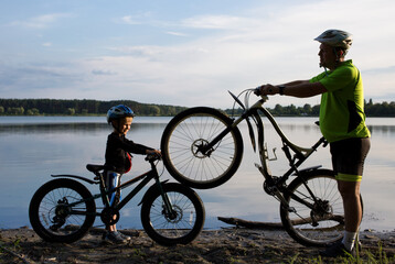 Silhouettes of an adult man and a boy with two bicycles in helmets near the lake at sunset. cycling, healthy lifestyle. Be like dad. Family pastime, dad and son ride a bike