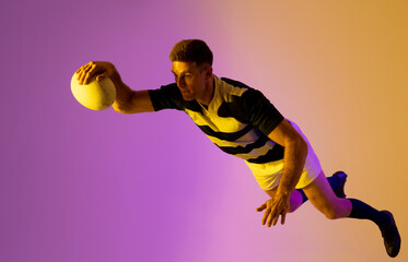 Caucasian male rugby player jumping with rugby ball over pink lighting