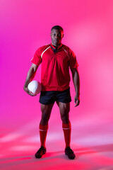 Portrait of african american male rugby player with rugby ball over pink lighting