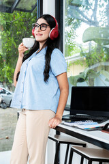 Asian woman with eyeglass listening to music with headphones and holding a cup of coffee with a laptop and a notebook with a mobile phone on the table