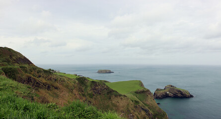 Cloudy weather on the green slopes of the coast of Northern Ireland over the idyllic island of Carrick-a-Rede with its narrow suspension bridge