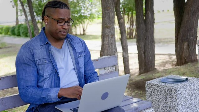 Young black man student does test work on laptop in park, takes off glasses from fatigue and rubs eyes. African-American man works online sitting in a park on a bench, yawning and looking at the clock