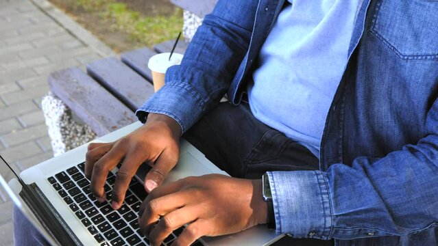 Close-up of the hands of a black student typing quickly on the keyboard of a laptop sitting in a park on a bench. African man typing a test paper, drinking coffee from a paper cup