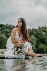 Fototapeta na wymiar Caucasian girl in a white dress with folded legs sits on the birch of the river and trims in the hands of entwined wreaths with polovy kvіtiv. The woman is guessing at the holy Midsummer