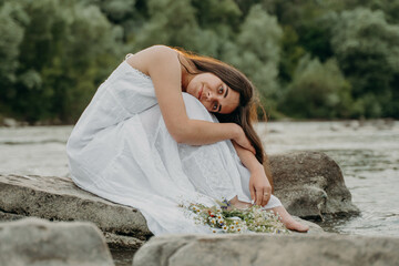 Attractive girl sits on stone with arms around knees and bowed head, with wreath of herbs and wildflowers next to her. Man enjoys nature. Resting by river with running water. Midsummer holiday