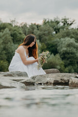 Fototapeta na wymiar Caucasian girl in a white dress with folded legs sits on the birch of the river and trims in the hands of entwined wreaths with polovy kvіtiv. The woman is guessing at the holy Midsummer