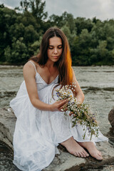 Beautiful Caucasian girl in white long dress sits on stones in middle of river and holds wreath of wildflowers in hands and looks at it. Cloudy gray sky. Woman in middle of nature. Midsummer holiday