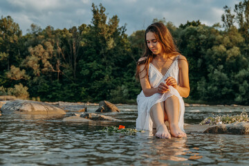 A cute girl sits on rock in middle of shallow river at sunset and releases a wreath of wildflowers and a candle into the water. Divination on day of midsummer on feast of Ivan Kupala. Pagan traditions