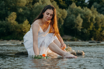 A cute girl sits on rock in middle of shallow river at sunset and releases a wreath of wildflowers and a candle into the water. Divination on day of midsummer on feast of Ivan Kupala. Pagan traditions