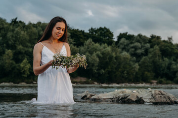 Fototapeta na wymiar Pretty girl in dress stands in middle of river in waist-deep water at sunset and releases wreath of wildflowers and candle into water. Magical divination on day of midsummer on feast of Ivan Kupala