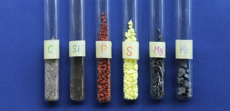 Simple inorganic chemicals in test tubes on a blue background: gray carbon, amorphous black silicon, crystals of red phosphorus, yellow sulfur, shavings of light magnesium, pieces of heavy lead.