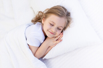 a cute baby girl hugs a pillow with her hands folded under her cheek and sleeps on the bed on a white cotton bed under a blanket, a healthy baby's sleep at night