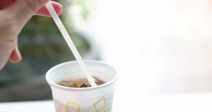 Hand holding straw and siring ice cola carbonate soft drink in white plastic glass.