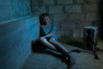A young dirty man in tattered clothes is locked in a concrete basement
