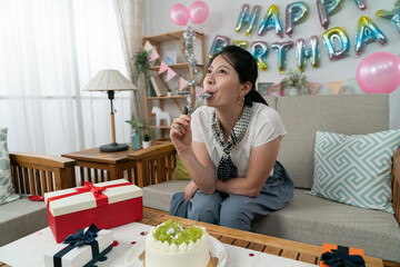 happy asian female licking cream and keeping spoon in mouth while enjoying tasty cake by herself at her birthday party in the apartment with celebration decors. - obrazy, fototapety, plakaty