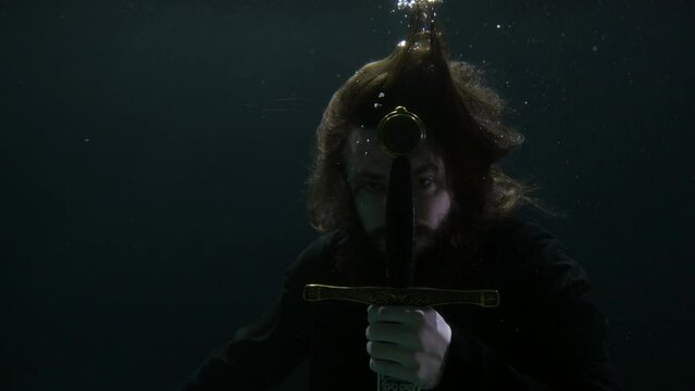 portrait of a long-haired man in dark water. he holds the hilt of a sword in front of face
