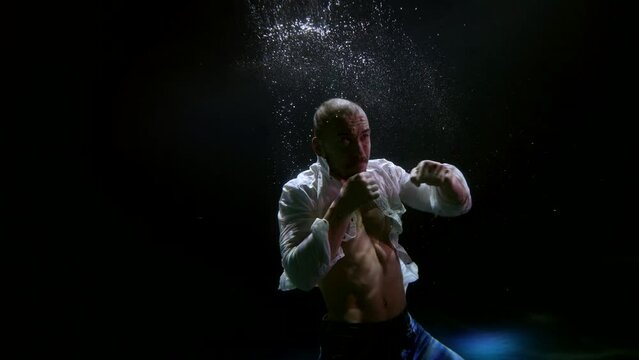 a man in an unbuttoned white shirt fights with a shadow in a stream of light with bubbles in the dark under water