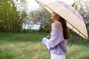 A girl in a sweater is standing under an umbrella. it's raining