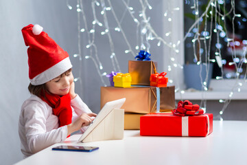 Winsome Caucasian Child Kid Girl In Santa Hat Using Laptop for Online Gift Present Search While Using Video Call Chat During Holidays At Home.
