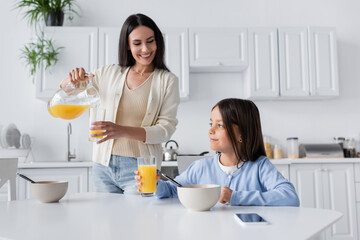 Obraz na płótnie Canvas brunette nanny pouring orange juice near smiling girl having breakfast in kitchen.