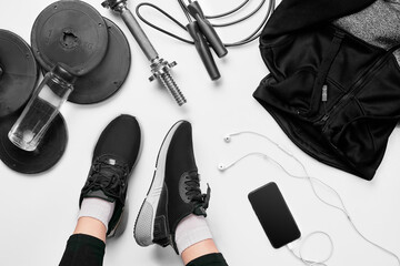 Women's legs in sneakers, fitness clothing, smartphone, dumbbells, headphones, top view.