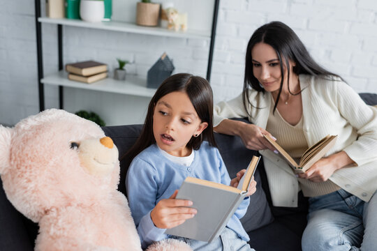 kid reading book to soft toy near smiling babysitter at home.