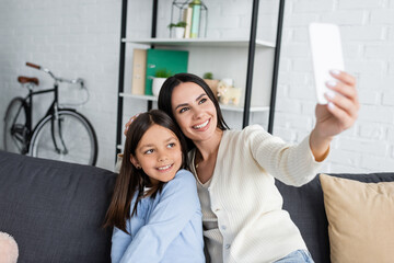 Fototapeta premium smiling babysitter taking selfie on cellphone with girl on sofa at home.