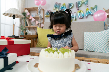 innocent asian baby girl looking at delicious cream cake on table during her second birthday celebration at home with colorful party decoration at background