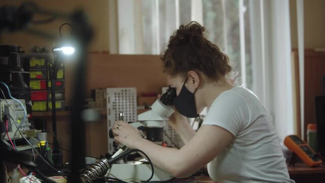Female worker at a service center using microscope and to electronics circiuit board. Girl looking through a microscope in workshop. Women and technical occupations.