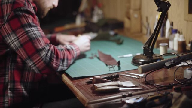 Close-up hands carve with the knife the pattern of leather and remove excess part. Tanner, tailor master works behind the workbench and creating leather goods. Handmade.