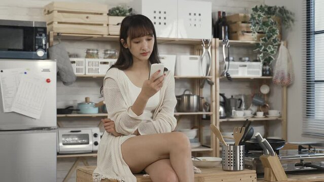 portrait of attractive asian female sitting cross legged on kitchen table is reading online articles on her smartphone with concentration during daytime at home