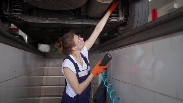 Female mechanic inspecting car suspension on hydraulic wheel play detector with remote control. Strong woman checks for wear in running gear and steering components. Service operator in overalls.