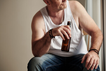 Cropped image of mature man in white undershirt drinking beer and looking outside through window