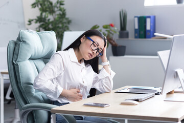 Abdominal pain at the workplace. A young beautiful Asian woman in glasses works in the office at...
