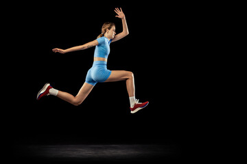 Fototapeta na wymiar Young sportive girl, long jumper in sports blue uniform performs triple jump isolated on black background. Concept of sport, energy, achievements, motion, speed.