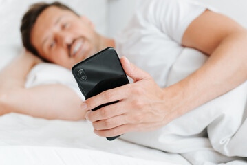 Close-up of happy man holding smartphone while lying in bed in early morning. Pleased guy using mobile phone in bedroom, looking at screen. Selective focus on device