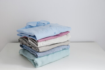 Stack of perfectly folded clothing items. Pile of different cold color shirts, sweaters and pants on the table, white wall background. Close up, copy space, studio shot.