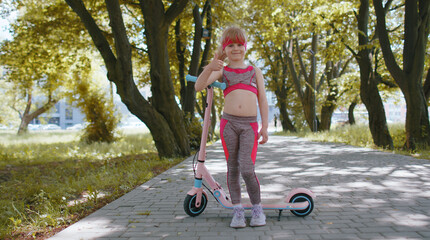 Athletic fit young girl rides on electric scooter on road in park on sunny day, outdoors. Child going standing near bike showing thumb up. Kid leisure activity, sport. Modern technology for children