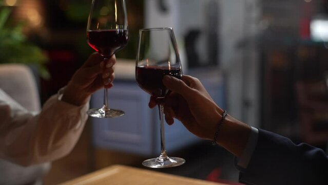 Close-up unrecognizable female and male hands clinking glasses with red wine and then drinking. Happy couple enjoying nice romantic dinner in fancy restaurant at evening. Shooting in slow motion.