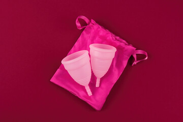 Two eco-friendly, reusable pink menstrual cups made of soft medical grade silicone rest on a silk storage pouch.  On a dark red blood-colored background