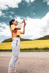 girl on the road, in a yellow T-shirt, taking a photo in a flowering field, against the backdrop of mountains