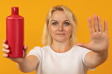 blonde woman showing stop sign with palm and holding shampoo bottle in hand against color background