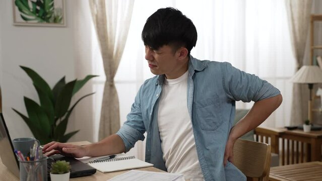 side view of an overworked asian male entrepreneur working from home is suffering backache and pounding massaging his lower back with a sigh in the living room