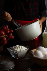 cook with apron whisking in a bowl with ingredients on the table, eggs flour bottle of milk and fruits