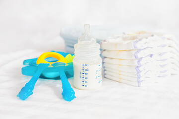 Fototapeta na wymiar Set of baby plastic dishes. milk bottle, feeding nibbler, blue cutlery, spoon, fork, diapers, toy, disposable diapers on the white bed background