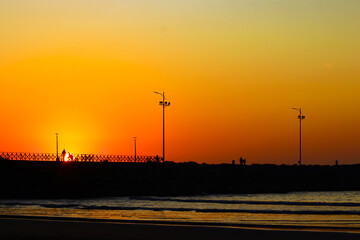 Fototapeta na wymiar Nascer do sol em Itajaí, Brasil, com céu laranja e amarelo refletindo no mar com o farol ao fundo e silhueta de pessoas. 